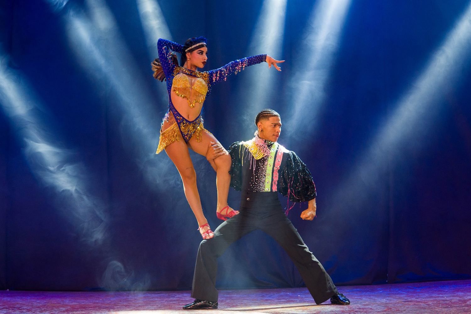 A dancer in blue and gold poised on her partner's thigh under hard white beams